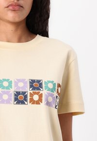 Geel katoenen T-shirt met een retro bloemenprint in blauw, groen, paars en oranje bloemenpatronen over de borst.