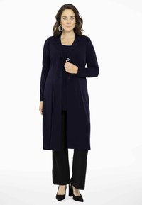 Marineblauwe lange cardigan met een kraag, knopen aan de voorkant, zijzakken, gecombineerd met een zwart top en broek. Gladde stof met een losse pasvorm.