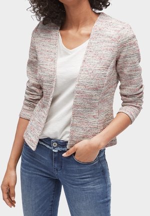 Kobieta ubrana w lekko szaroróżowy, teksturowany, otwarty żakiet na biały T-shirt z dekoltem w serek i niebieskie jeansy, z ręką w kieszeni.