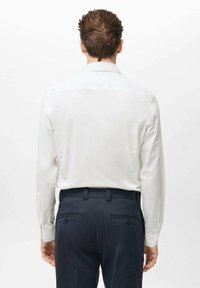 Camisa blanca de manga larga con cuello clásico y puños abotonados, combinada con pantalones de sastre azul oscuro, vista desde atrás.