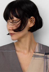 Gros plan sur une personne aux cheveux noirs et à la coupe bob, portant une veste grise texturée avec des motifs abstraits clairs et des rayures.