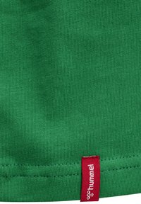 Zielona bawełniana tkanina o ribbowanej teksturze, z czerwoną metką z napisem "hummel" przy dole. Prosty design bez dodatkowych wzorów.