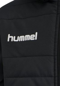 Černá zateplená bunda s hladkou texturou, se zvýrazněnými horizontálními švy a výrazným bílým logem "hummel" na hrudi.