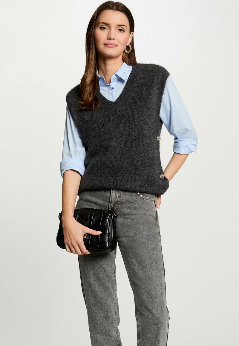 Gilet di lana grigio scuro con scollo a V, indossato sopra una camicia azzurra chiara. Borsa a tracolla nera trapuntata tenuta in mano dalla modella, abbinata a jeans grigi.