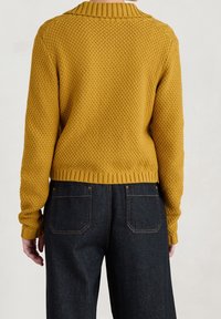 Kvinna som bär en senapsgul texturerad stickad tröja och mörkblå denimjeans, visad från baksidan mot en enfärgad bakgrund.