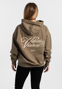 Hnedá mikina s kapucňou s oversized strihom, textúrovaným povrchom a bielym potlačeným textom "Hodnoty a vízie" na chrbte.
