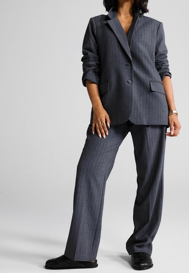 Femme portant un costume gris foncé à fines rayures avec un blazer et un pantalon à jambes larges, associé à des chaussures plates noires sur un fond clair.