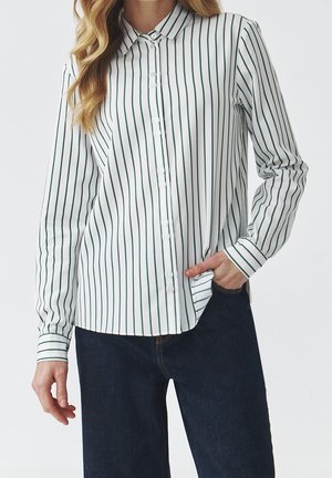 Kvinde iført en hvid langærmet skjorte med tynde lodrette sorte striber og mørkeblå jeans, hånd i lommen.
