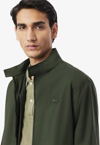 Giacca verde con zip e collo alto, con logo discreto sul lato sinistro del petto, abbinata a una polo verde chiaro sotto. Tessuto liscio e texture.