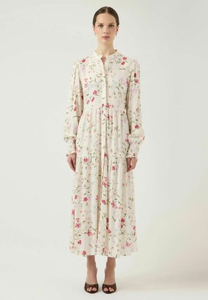 Kvinde iført en langærmet, ankel-længde hvid kjole med et pink og grønt blomsterprint og brune åbne hæle, stående på en ensfarvet baggrund.