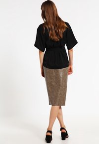 Blouse noire à manches larges, taille cintrée, associée à une jupe texturée marron clair mi-longue avec un motif subtil. Sandales noires.