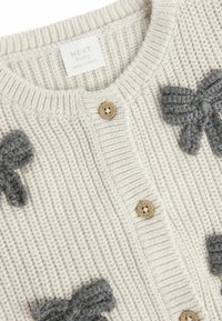 Kremīgi adīta zīdaiņu kardigāna, kas izceļas ar pelēkām adītām loku akcentēm, ribu faktūru un koka pogām aizdarei. Uz etiķetes rakstīts "NEXT baby".