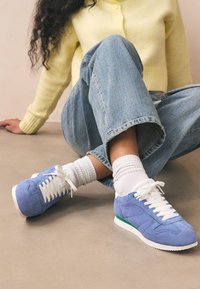 Sneakers in pelle scamosciata blu con lacci bianchi, caratterizzate da un accento verde sulla suola. Abbinate a denim comodi e calzini bianchi.