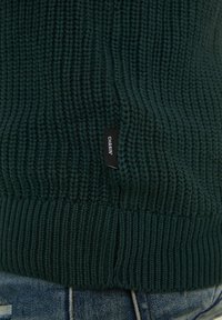 Tmavě zelený pletený svetr s žebrovanou texturou a štítkem na lemu označeným "CHASIN'." Tkanina má silný, útulný vzhled.