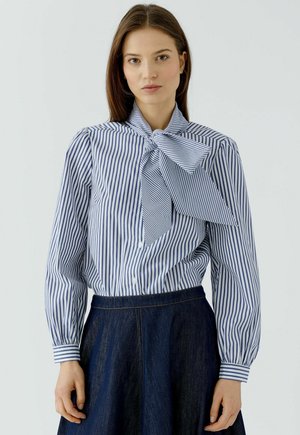 Vrouw die een blauw-wit verticaal gestreept blouse draagt met een grote strik bij de hals en een donkerblauwe rok, staande tegen een effen achtergrond.