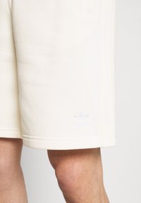 Krämsfärgade bomullsshorts med en slät yta, sidofickor och en broderad ljuslila Adidas-logotyp nära fållen.