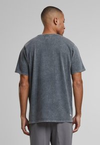 Szary, wyblakły t-shirt bawełniany o luźnym kroju, z okrągłym dekoltem i krótkimi rękawami. Widoczne szwy i małe logo na dolnym brzegu.