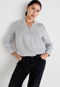 Bela in mornarsko modra slečena bluzica s henley ovratnikom, dolgimi rokavi in gumbnimi detajli, v kombinaciji s črnimi kavbojkami. Gladka tekstura.