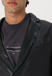 Schwarze Lederblazer mit einer einzelnen Brusttasche, über einem dunkelgrauen Sweatshirt mit weißen Buchstaben getragen. Glatte Textur und taillierte Passform.