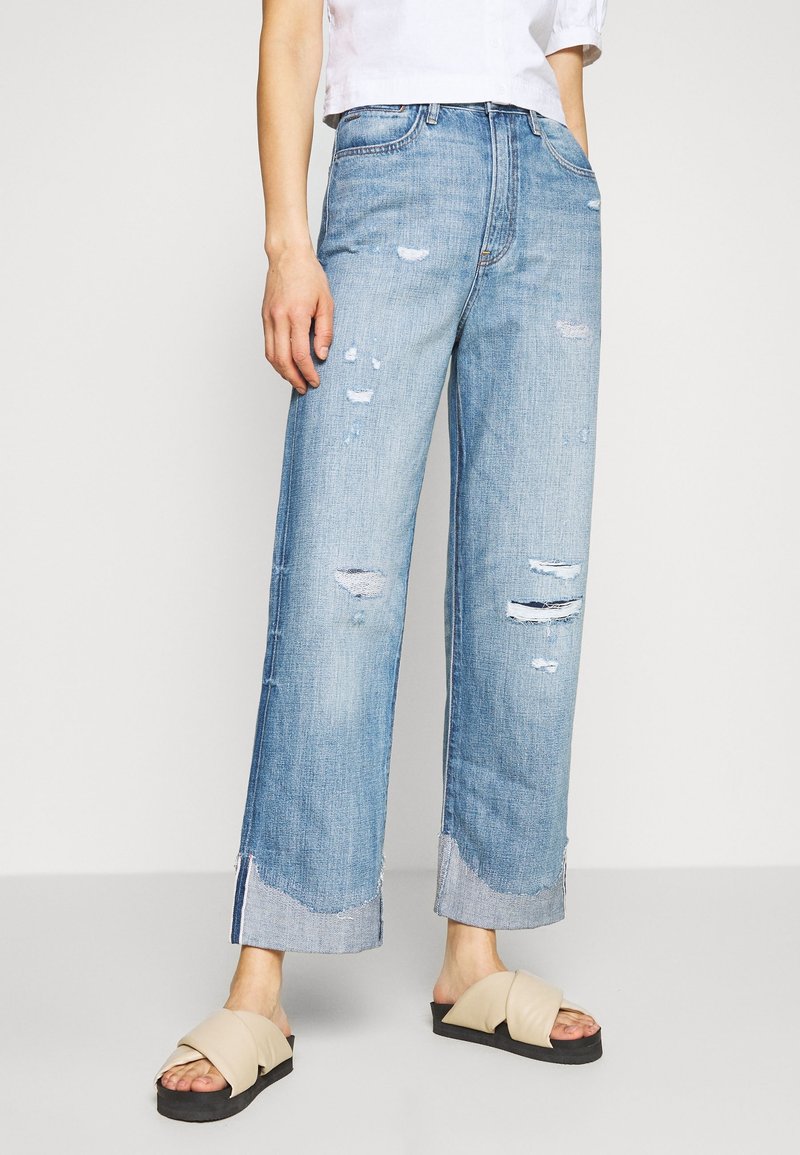 Jeans de pierna ancha de un azul claro desgastado, con desgastes y deshilachados en el dobladillo. Llevados con sandalias deslizantes beige. La tela muestra una textura clásica de mezclilla.