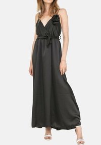 Robe maxi noire sans manches avec un décolleté en V, un détail floral sur le côté, une ceinture à nouer à la taille et une texture satinée lisse. Portée avec des sandales à talons blanches.