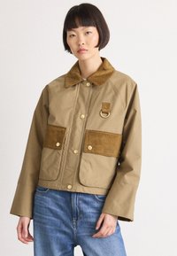 Bruine jas met een corduroy kraag en zakken, voorzien van goudkleurige hardware en een gestructureerde stof, gecombineerd met lichtblauwe jeans.