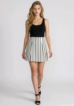 Mujer joven con cabello largo y rubio, vestida con un top negro sin mangas, falda blanca con rayas verticales negras, y sandalias negras de tacón alto.