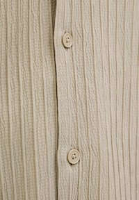 Beige gestructureerde stof met verticale strepen, in het midden vastgemaakt met een ronde, lichtgekleurde knoop.