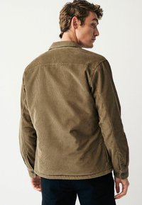 Brown corduroy jacket with a relaxed fit, featuring vertical ribbing, a pointed collar, and buttoned cuffs. Rear view, plain background.
