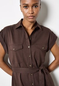 Robe chemise marron à manches courtes, deux poches avant, fermeture à boutons et une ceinture assortie. Texture lisse avec un léger lustre.