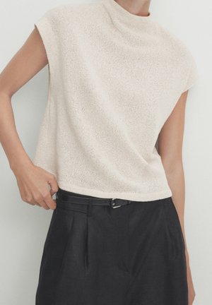 Personne portant un haut sans manches beige texturé, rentré dans un pantalon noir plissé à taille haute avec une ceinture noire fine.