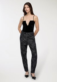 Kobieta stoi z rękami na plecach, ubrana w czarną aksamitną koszulę z głębokim dekoltem w kształcie V, ciemne dżinsy w wysokim talii w kwiatowy wzór oraz czarne szpilki.