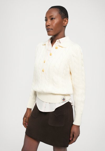 Pull en tricot crème avec un motif en cable et des boutons jaunes, porté par-dessus une chemise à col rose pâle. Associé à une mini-jupe en daim marron avec deux poches.