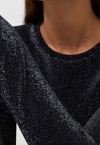 Top nero a maniche lunghe in tessuto testurizzato scintillante con scollatura rotonda, che mostra un primo piano della manica e della spalla.
