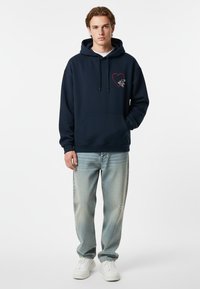 Felpa con cappuccio blu navy con grafica a cuore rosso e dettagli grafici. Abbinata a jeans chiari strappati e sneakers bianche.