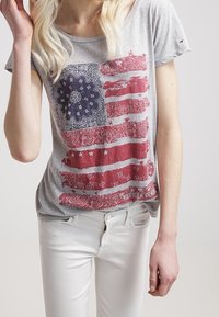 Gray short-sleeve T-shirt featuring a red and blue American flag design with paisley patterns and white stars, paired with white jeans.