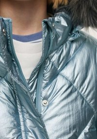 Padded, metalická modrá bunda s prošívaným vzorem, na zip a s kapucí. Obsahuje stříbrné patentky a kontrastní vnitřní vrstvu.