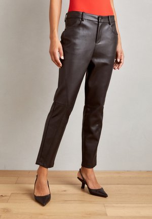 Frau in schwarzen Lederhosen und schwarzen spitzen Slingback-Highheels, die auf einem Holzfußboden vor einer schlichten Wand steht.