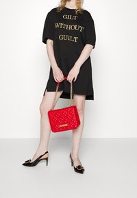 Černé oversized tričko se zlatým textem, červená prošívaná kabelka s zlatým řetízkem a černé lodičky na přezku s zlatými akcenty.