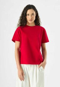Rotes Baumwoll-T-Shirt mit rundem Halsausschnitt und kurzen Ärmeln. Glatte Textur, gerader Schnitt, mit einem kleinen Logo auf der Brust.