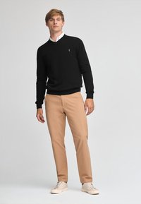 Suéter negro con escote en v y logo, combinado con pantalones beige y zapatillas ligeras. El conjunto presenta un ajuste relajado y una textura de punto.
