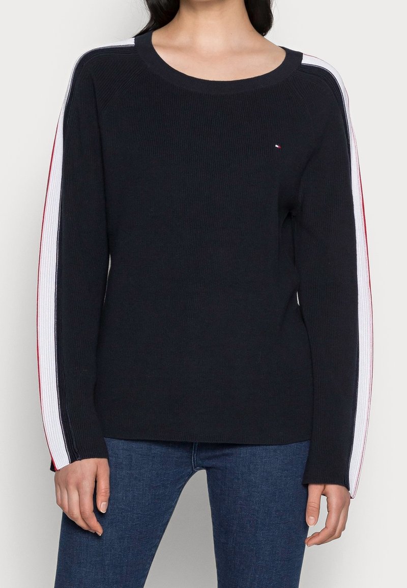 Femme portant un pull noir à manches longues avec des rayures blanches et rouges le long des manches et un jean bleu, debout devant un fond uni.