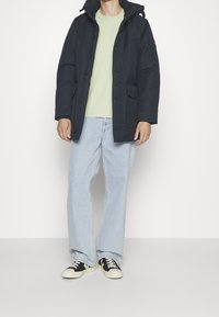 Tummansininen toppatakki hupulla, jossa vetoketju ja nepparikiinnitys, rintatasku, päällä vaaleanvihreä t-paita ja vaaleat farkut.