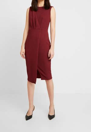Vrouw draagt een mouwloze bordeauxrode jurk tot op de knie met een asymmetrische vooraanstaande split en zwarte puntige hakken, staand tegen een effen achtergrond.