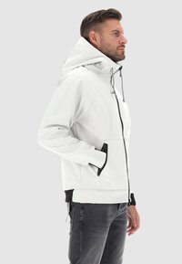 Giacca bianca con cappuccio, dal tessuto liscio, con tasche con zip e dettagli neri sui polsini e sull'orlo; presenta una vestibilità rilassata e un cappuccio regolabile.