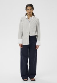 Jeune femme aux longs cheveux portant une chemise gris clair à boutons avec des poignets blancs et un pantalon large foncé, posant devant un fond uni.