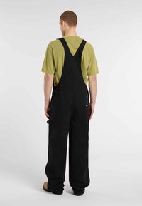 Salopette en coton noir avec jambes larges, bretelles ajustables et plusieurs poches, associée à une chemise à manches courtes verte.