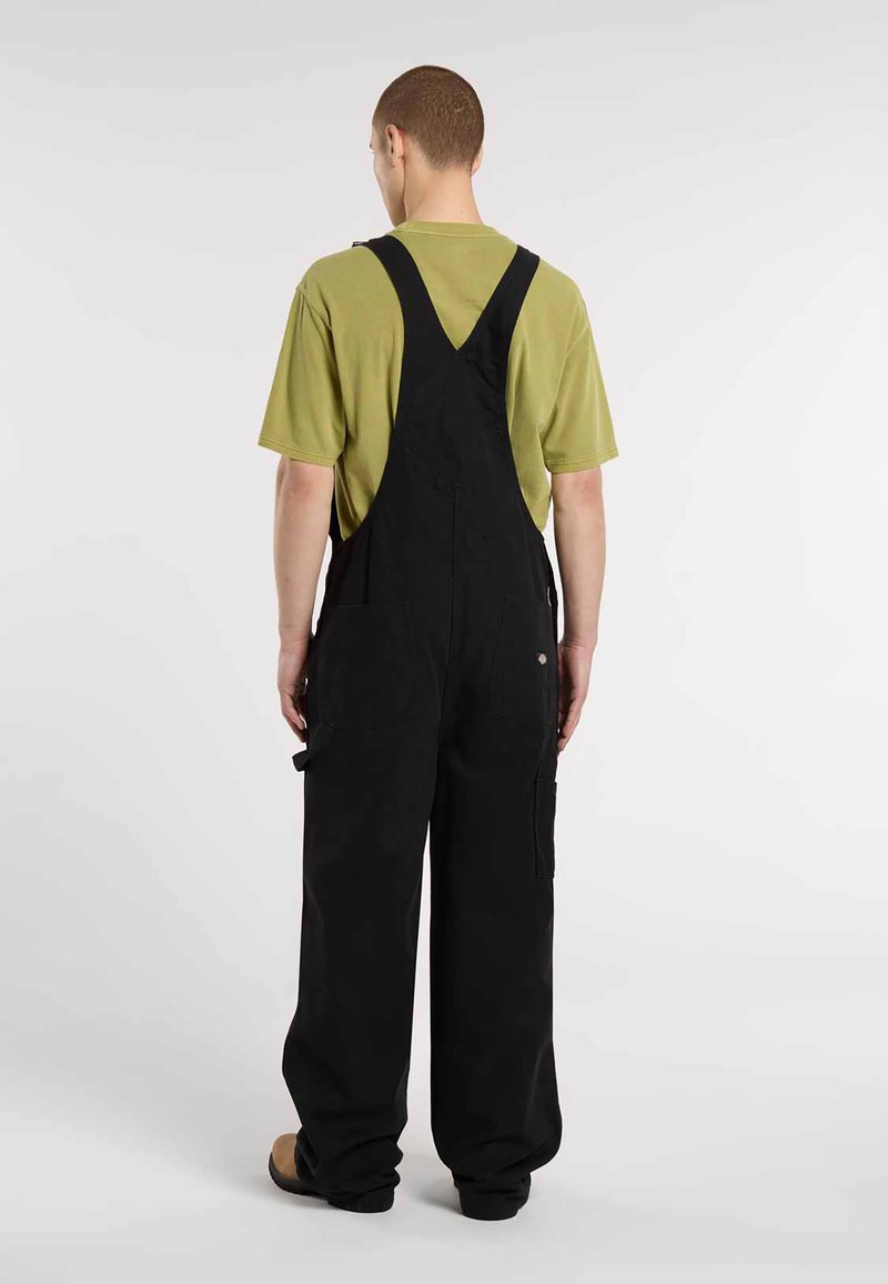 Sort bomuldsoveralls med vide ben, justerbare stropper og flere lommer, parret med en grøn kortærmet skjorte.