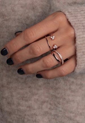 Tres anillos en una mano: una delgada banda de oro rosa, un anillo curvado de oro rosa y un anillo de oro rosa con pequeñas piedras preciosas transparentes.