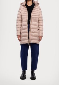 Mujer vestida con abrigo acolchado beige con capucha, camisa negra, pantalones azules y botas negras gruesas, de pie contra un fondo blanco.